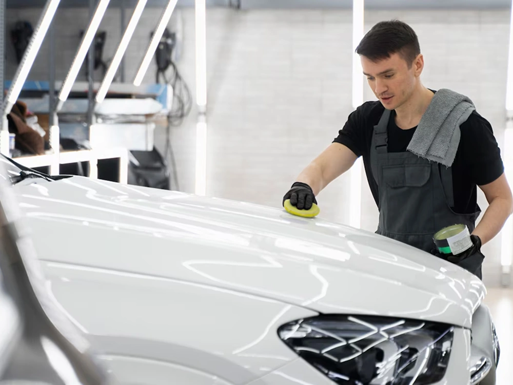 Auto detailer in overalls applies wax or polish to the hood of a white car using a yellow applicator pad in a well-lit garage, with a towel draped over his shoulder and a container of product in hand.