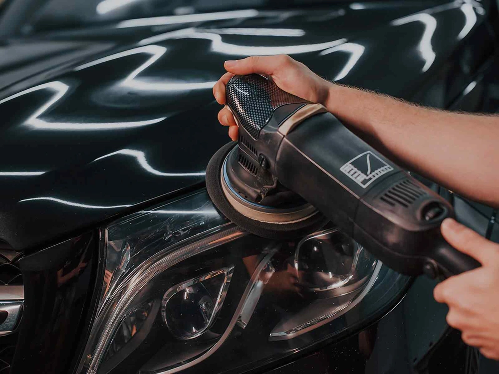 A person uses a dual-action polisher on the hood of a black car near the headlight, creating a glossy reflection as part of a paint correction or detailing process.
