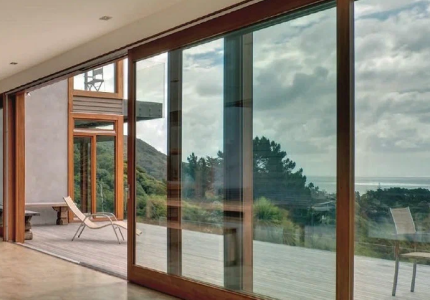 Large floor-to-ceiling sliding glass doors with wooden frames open to a deck overlooking a scenic view of hills, trees, and the ocean under a cloudy sky. The indoor-outdoor transition emphasizes modern architecture and natural surroundings.