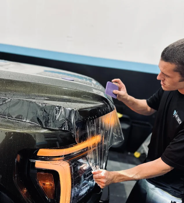 A technician applies a clear paint protection film (PPF) to the headlight and front fender of a truck, using a squeegee to smooth the film over the surface under bright shop lighting.