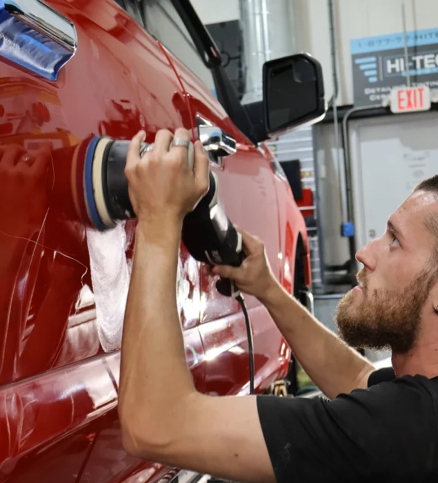 A technician uses a dual-action polisher to buff the side of a red truck inside the Hi-Tech Appearances workshop.