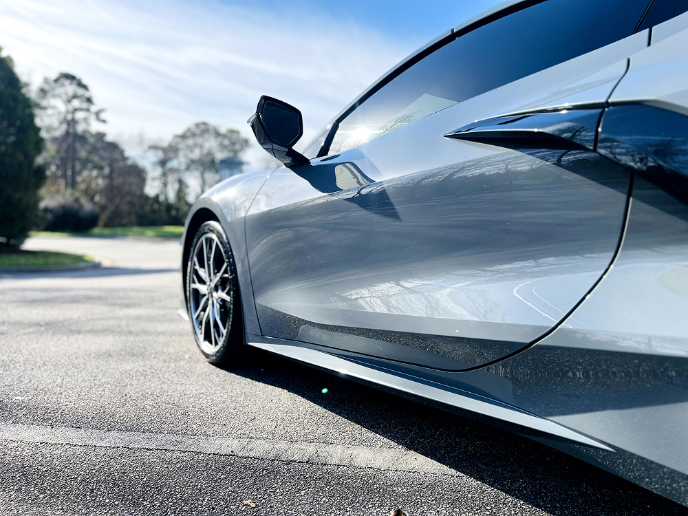 Close-up side view of a sleek, silver sports car parked on a sunlit road, showcasing its aerodynamic curves, glossy finish, and black alloy wheels with low-profile tires. The car reflects the surrounding trees and sky on its polished surface.