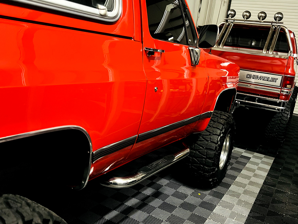 Two vintage red Chevrolet trucks with lifted suspensions and oversized tires parked indoors on a checkered garage floor, with one showcasing a chrome roll bar and off-road lights on the rear.