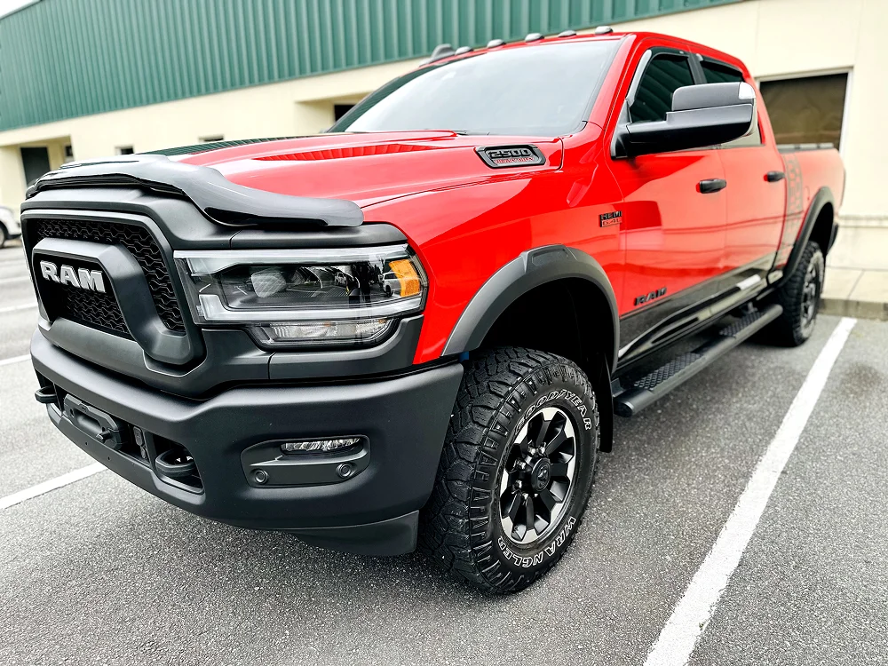 Bold front-side view of a red RAM 2500 heavy-duty pickup truck with blacked-out trim, rugged off-road tires, a vented hood, and “HEMI 6.4L” and “2500 Heavy Duty” badging. The truck is parked in a lot outside a commercial building.