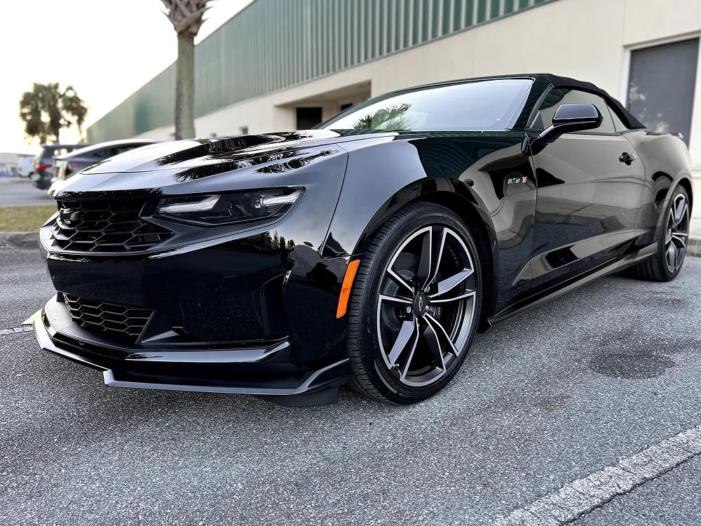 Glossy black Chevrolet Camaro convertible parked in a lot, captured from the front-side angle with the top up, featuring sharp headlights, bold front grille, and five-spoke wheels.