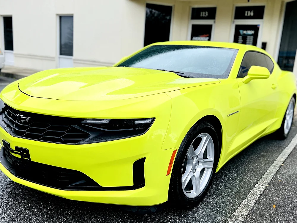 A bright neon yellow Chevrolet Camaro parked outside a building with rain droplets on the hood, showcasing its sleek design and glossy paint finish.