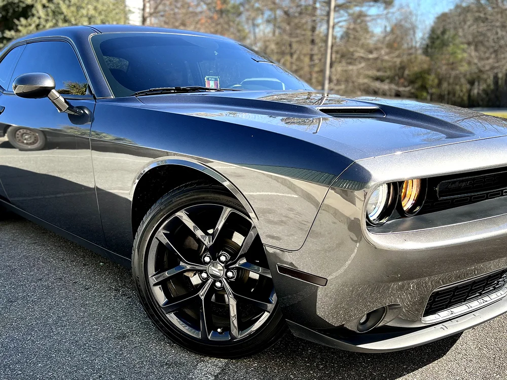 Front-side view of a dark gray Dodge Challenger parked in daylight, featuring aggressive styling, gloss black wheels, a hood scoop, and one headlight turned on. The car’s reflective surface mirrors the surrounding environment.
