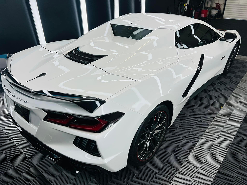 Rear and side angle of a white Chevrolet Corvette in a showroom, showcasing its bold rear spoiler, black accents, sculpted body lines, and signature tail lights. The car is parked on a patterned garage floor under bright studio lighting.