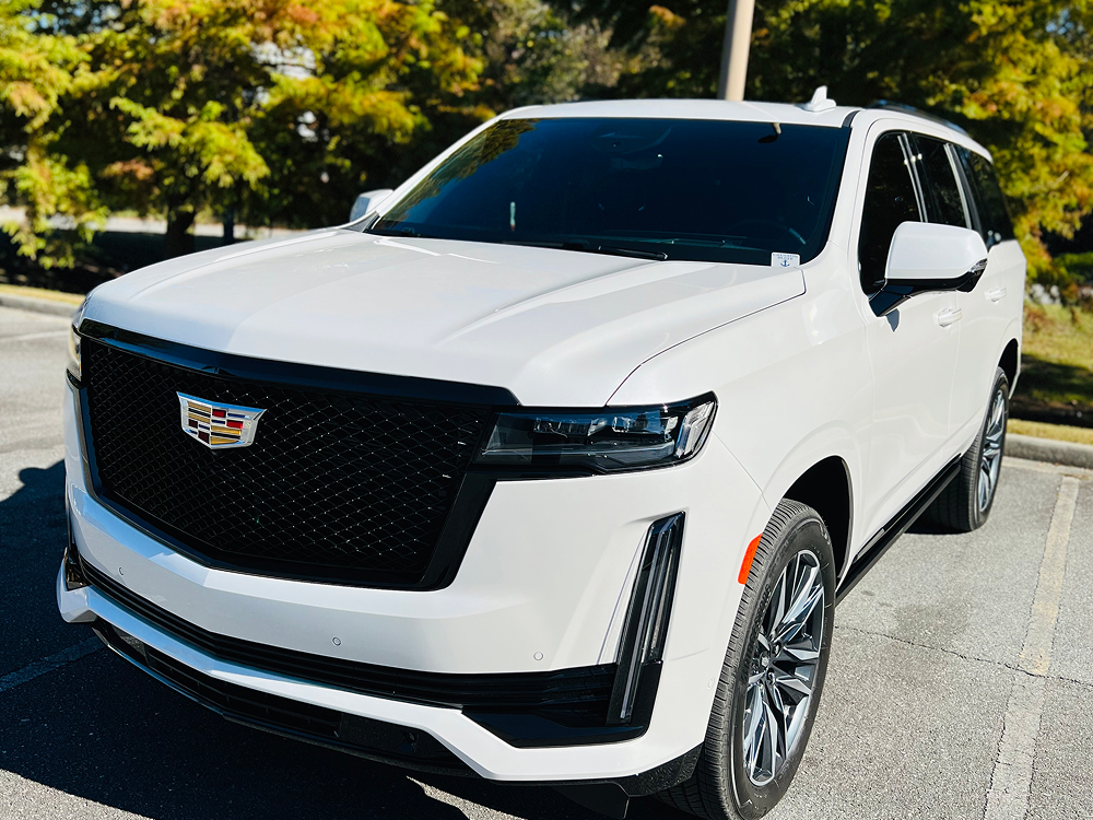 Front three-quarter view of a white Cadillac Escalade parked outdoors, featuring a bold black grille with the Cadillac emblem, sleek vertical LED headlights, and large chrome wheels. The SUV is positioned in a sunny parking lot with trees in the background.