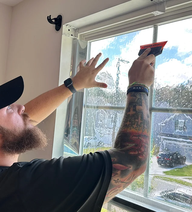 Bearded man with a tattooed arm installs window film by smoothing it onto a wet residential window using a red squeegee tool, with sunlight streaming through the glass.