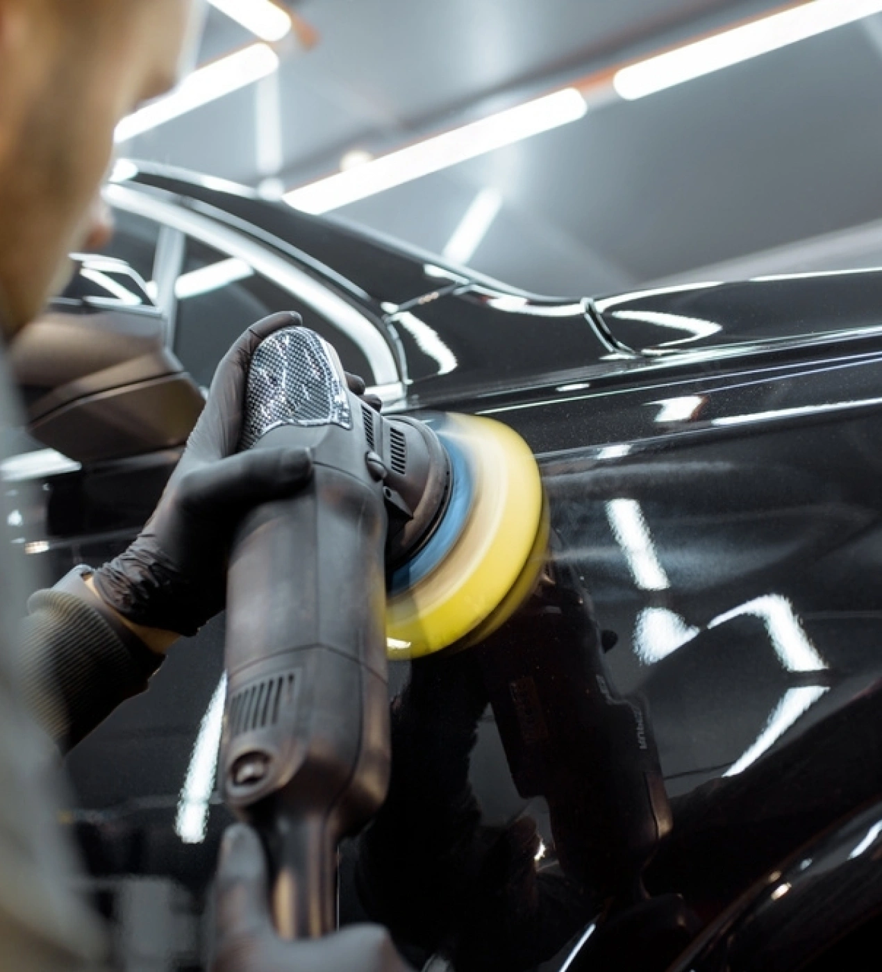 Auto detailer using a dual-action polisher with a yellow foam pad to buff the black exterior of a car, enhancing shine and removing imperfections under bright shop lighting.