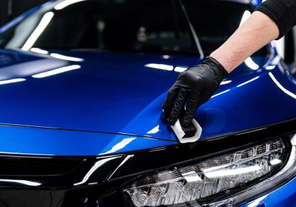 Auto detailer wearing a black glove applies ceramic coating to the hood of a shiny blue car using an applicator pad, enhancing gloss and protection.