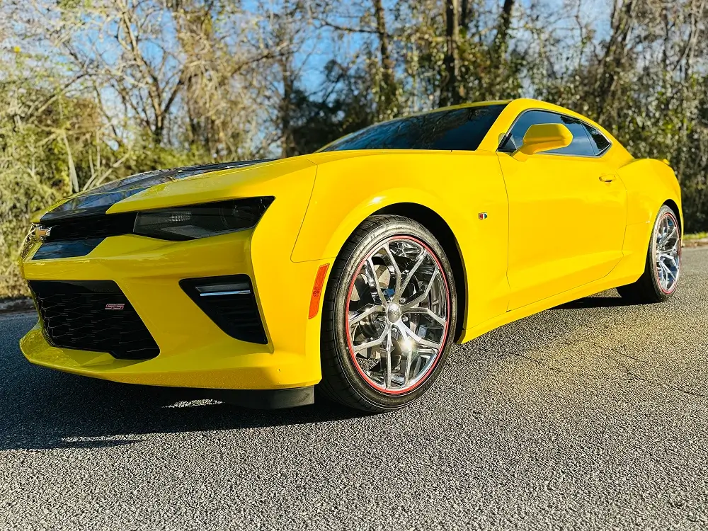Bright yellow Chevrolet Camaro SS parked on a sunlit road, viewed from a low front-side angle, featuring chrome rims with red trim, blacked-out front grille, and aggressive body lines.