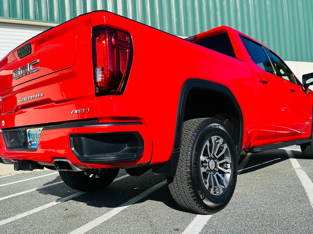 Bold red GMC Sierra AT4 truck photographed from a low rear-side angle, showcasing its lifted stance, aggressive off-road tires, dual exhaust, and large GMC tailgate branding.