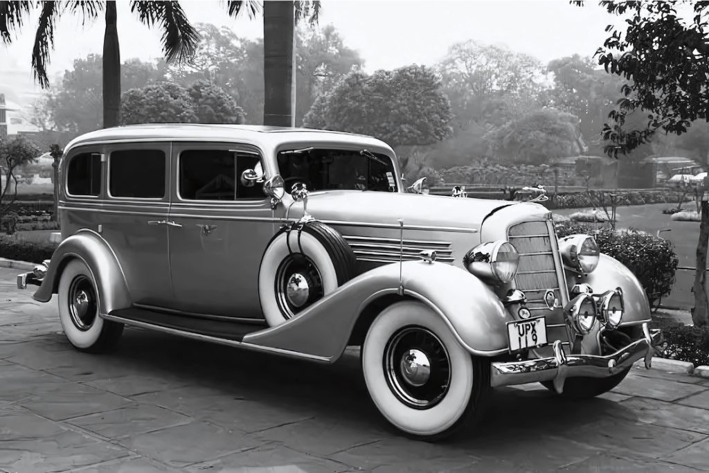 A vintage luxury car from the 1930s with whitewall tires, side-mounted spare wheels, and a chrome front grille is parked on a stone driveway surrounded by palm trees and manicured gardens. The black-and-white photo captures the era before modern window tinting became common.