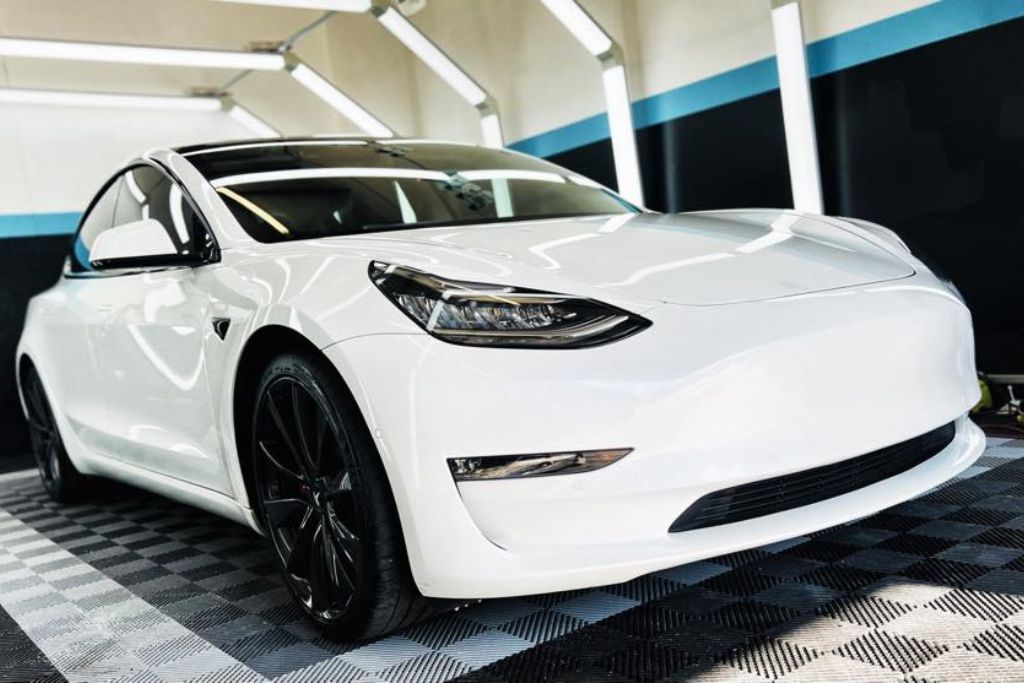 A white Tesla with black wheels sits under bright lights in a detailing bay, showcasing a flawless glossy finish likely protected by full-body paint protection film. The image reflects the premium quality of professional PPF application.