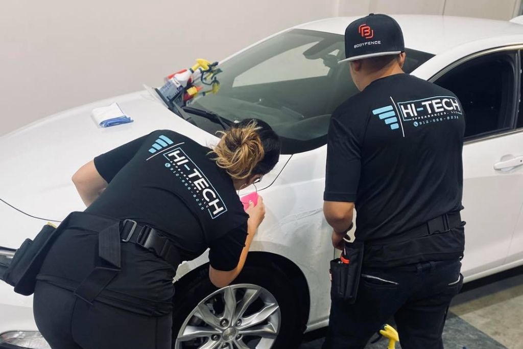 Two Hi-Tech Auto Spa technicians apply paint protection film to the front fender of a white car, carefully smoothing the film for a seamless finish. Both wear branded uniforms, emphasizing professional installation and attention to detail.