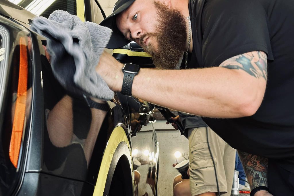 A focused auto detailer in a black shirt and cap polishes a car’s exterior with a gray microfiber cloth, reflecting his concentrated expression in the glossy black paint. Yellow tape outlines the car panel for precision during the detailing process.