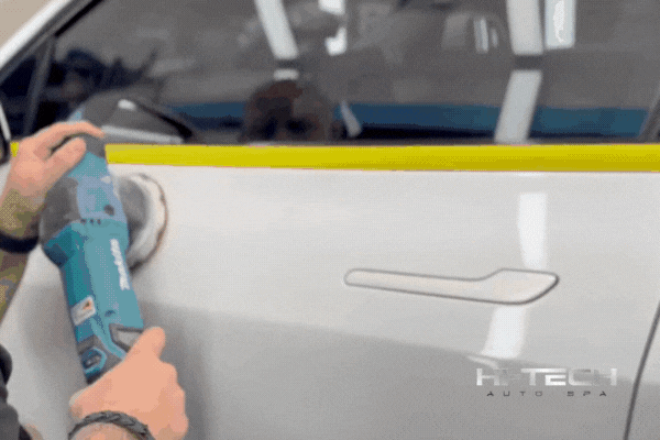 A technician uses a power polisher on the side of a white car with yellow masking tape along the edge, preparing the surface for ceramic coating at Hi-Tech Auto Spa. This visual highlights the importance of expert prep in regions like Savannah, where ceramic coating helps defend against coastal elements.