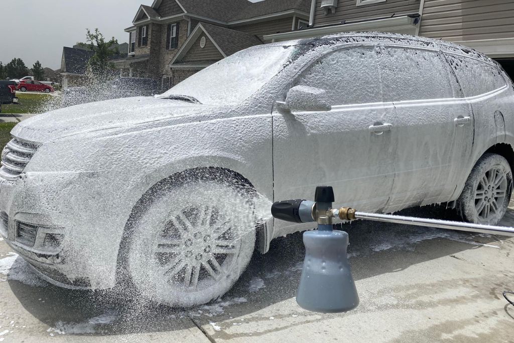A white SUV is being coated in thick foam using a pressure washer with a foam cannon attachment, demonstrating proper handwashing techniques for maintaining a ceramic-coated vehicle during summer.