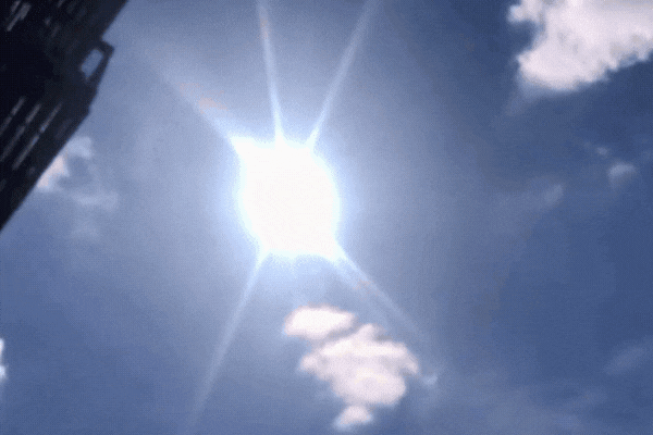 A bright summer sun blazes in a mostly clear blue sky over Savannah, Georgia, with a few scattered clouds and a building partially visible on the left, illustrating the intense UV exposure typical of Savannah's climate.