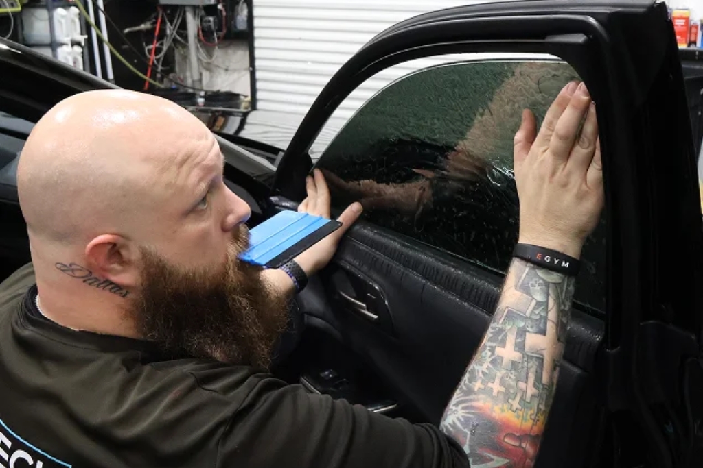 A tattooed technician carefully applies window tint film to a car door using a squeegee tool, ensuring a smooth finish. The image highlights the precision and professionalism involved in installing high-performance window tint.