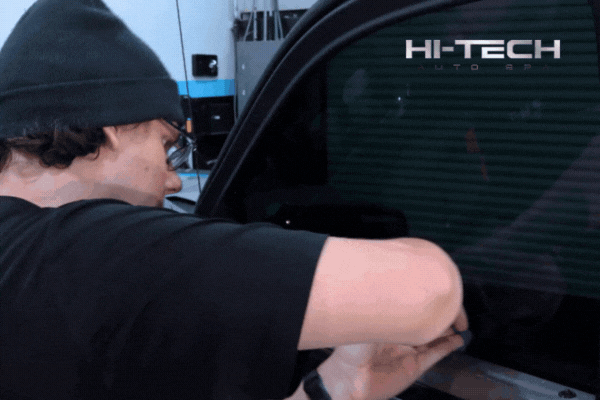 A technician wearing a beanie and glasses installs window tint film on a car door inside a professional shop, with “HI-TECH AUTO SPA” branding visible on the tinted window. The scene reflects expert-level service and attention to detail in a climate-controlled environment.